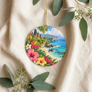 Puede incluir: Un adorno redondo de cerámica que presenta una vibrante escena costera tropical con un templo balinés en un acantilado, aguas oceánicas turquesas y una playa de arena, enmarcado por coloridas flores de hibisco rosas, amarillas y blancas.