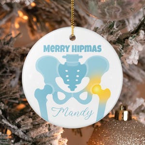 Puede incluir: Adorno navideño redondo y blanco con la inscripción "MERRY HIPMAS" en azul claro. Presenta una ilustración en azul claro de una pelvis y una articulación de la cadera resaltada en amarillo. El nombre "Mandy" está escrito debajo.