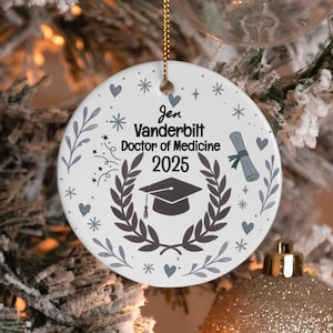May include: Round white ceramic graduation ornament with a gold hanging string. It features the text "Jen Vanderbilt Doctor of Medicine 2025", a mortarboard, laurel wreath, and other gray decorative elements.