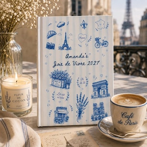 May include: A hardcover journal with light blue vertical stripes and blue illustrations of Paris landmarks, including the Eiffel Tower, Arc de Triomphe, and text reading Amanda's Joie de Vivre 2027, alongside a candle and a cup of coffee.
