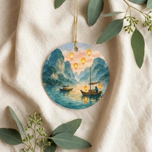Könnte beinhalten: Ein runder Keramikschmuck mit einer gemalten Landschaft aus einem ruhigen Gewässer, das von hohen, zerklüfteten Bergen umgeben ist. Zwei kleine Holzboote mit Masten schwimmen auf dem Wasser, beleuchtet von leuchtenden orangefarbenen Papierlaternen.