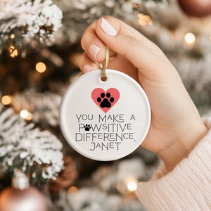 Op de afbeelding: Een witte keramische kerstbal met een gouden lint. De bal heeft een roze hart met een zwarte pootafdruk en de tekst "YOU MAKE A PAWSITIVE DIFFERENCE, JANET."