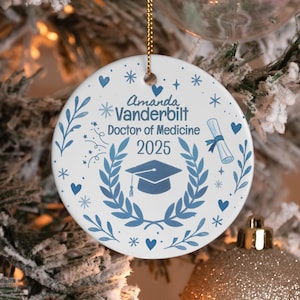 May include: A white ceramic Christmas bauble with blue detailing. The bauble displays the text "Amanda Vanderbilt Doctor of Medicine 2025" and a graduation cap. Decorative elements include hearts, stars, and laurel leaves.