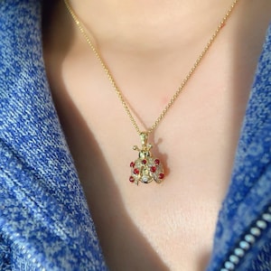 May include: A gold-colored necklace with a ladybug pendant. The ladybug pendant is adorned with red and clear gemstones. The necklace is displayed against a blue background.