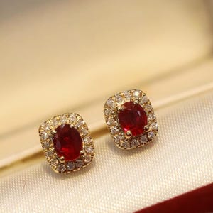Puede incluir: Un par de pendientes de botón con rubíes rojos de corte ovalado engastados en oro amarillo. Cada rubí está rodeado por un halo de pequeños diamantes blancos redondos y claros. Los pendientes tienen una forma rectangular clásica.