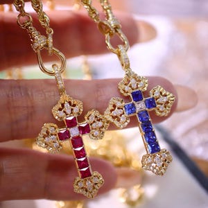 Pode incluir: Dois pingentes de cruz dourados com detalhes de pedras preciosas. Uma cruz apresenta pedras vermelhas, a outra azuis. Ambas têm enfeites semelhantes a diamantes e são suspensas por correntes de ouro. As cruzes são seguradas contra um fundo desfocado.