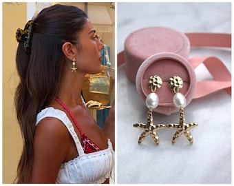 May include: Gold-colored starfish earrings with white pearl accents. The earrings are displayed in a pink velvet jewelry box. The earrings are hanging from a gold-colored stud with a textured design.