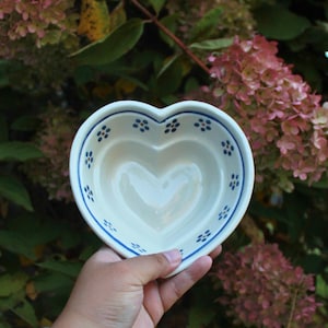 May include: A heart-shaped ceramic bowl with a white interior and a blue floral pattern around the rim. The bowl is held against a backdrop of green leaves and pink flowers.