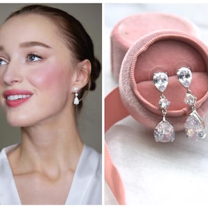 May include: Elegant silver drop earrings with clear, sparkling cubic zirconia stones. The earrings feature a teardrop design and are displayed in a pink velvet jewelry box. A woman is wearing the earrings in the other image.