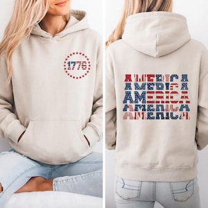 Könnte beinhalten: Ein beigefarbener Kapuzenpullover mit einer Vordertasche und einer Kapuze. Auf der Vorderseite ist das Jahr "1776" in einem roten und blauen Sternmuster abgebildet. Auf der Rückseite steht das Wort "AMERICA" in einem patriotischen rot-weiß-blauen Muster.
