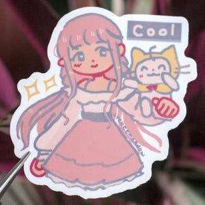 Puede incluir: Una pegatina con un personaje de pelo rosa con un gato que sostiene un cartel que dice "Cool".