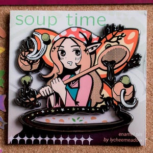 May include: Enamel pin featuring a cartoon character stirring a pot of soup. The pin has a black outline with orange, pink, and white accents. The text "soup time" is at the top, and "enamel pin by lycheemeadow" is at the bottom.