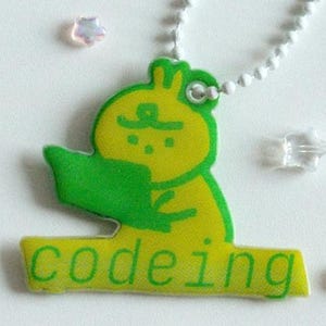 Puede incluir: Un llavero amarillo y verde con un personaje de dibujos animados leyendo un libro con la palabra "codeing" en verde. El llavero está sujeto a una cadena de bolas plateada. El fondo es blanco con decoraciones en forma de estrella dispersas.