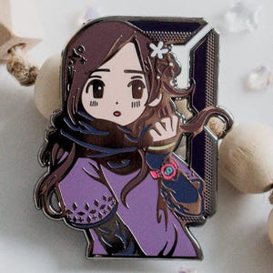 Puede incluir: Un pin metálico con una ilustración de dibujos animados de una mujer con cabello castaño y un atuendo morado. El pin está sobre un fondo blanco con cuentas de madera decorativas y cristales transparentes. El texto "lycheemeadow" está en la parte inferior.