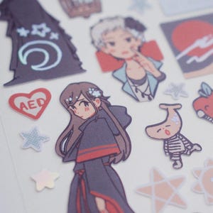 Puede incluir: Una hoja de pegatinas con ilustraciones de estilo anime. Las pegatinas presentan personajes, estrellas, un corazón con "AED" y otros elementos decorativos. La paleta de colores es suave, con tonos pastel y toques iridiscentes.