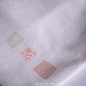 May include: White fabric tote bag with three pink and green floral designs. The designs are in the shape of squares and feature a swirl pattern.