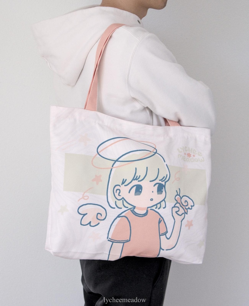 May include: A pink canvas tote bag featuring a cartoon illustration of a girl with blonde hair and a halo, holding a butterfly. The text "lychee meadow" is printed on the bottom of the bag.