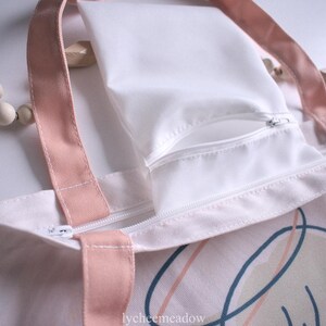 May include: A white canvas tote bag with pink straps and a white zippered pocket. The bag has a pink and white abstract design with a cartoon face on the front. The text "lycheemeadow" is printed on the bottom of the bag.