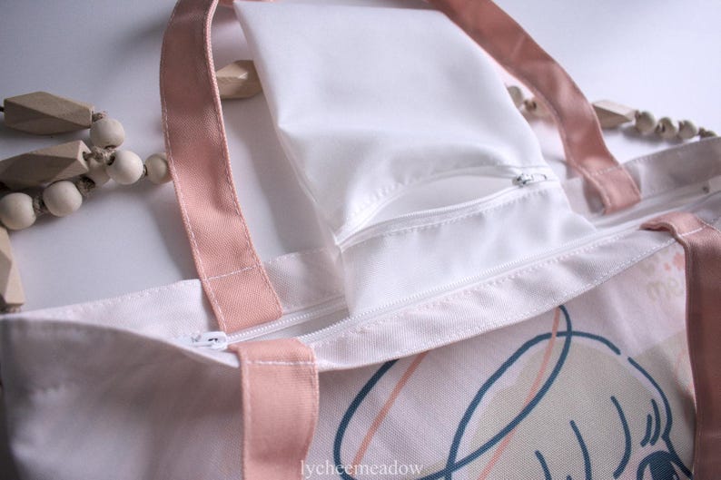 May include: A white canvas tote bag with pink straps and a white zippered pocket. The bag has a pink and white abstract design with a cartoon face on the front. The text "lycheemeadow" is printed on the bottom of the bag.