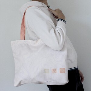 May include: A white tote bag with pink straps and a pink, white and green design. The bag has three small squares with different designs on it.