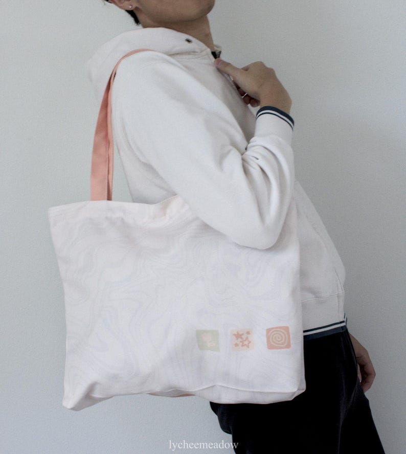 May include: A white tote bag with pink straps and a pink, white and green design. The bag has three small squares with different designs on it.