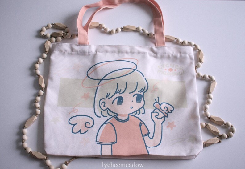 May include: A white tote bag with a pink handle and a cartoon illustration of a girl with a halo and wings. The girl is holding a butterfly and the text "lycheemeadow" is printed on the bottom of the bag.