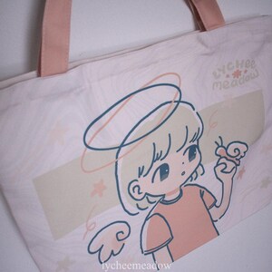 May include: A pink canvas tote bag featuring a cartoon illustration of a girl with a halo and wings. The girl is holding a butterfly and the bag has the text "lychee meadow" printed on it.