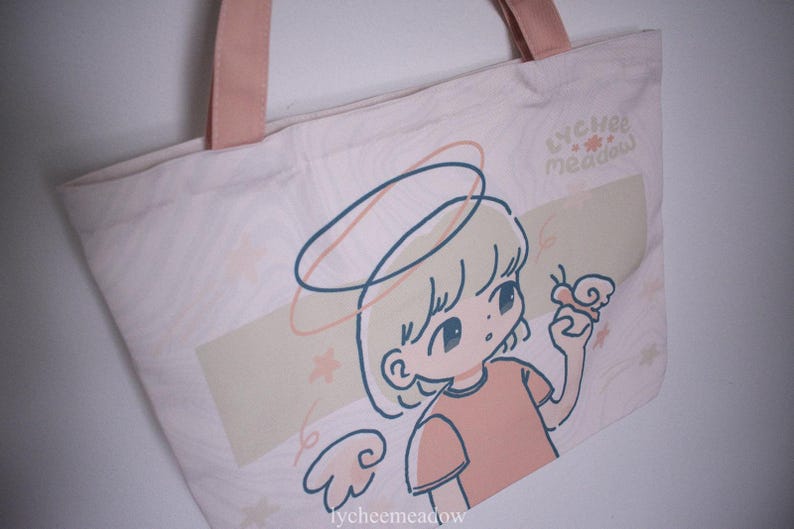 May include: A pink canvas tote bag featuring a cartoon illustration of a girl with a halo and wings. The girl is holding a butterfly and the bag has the text "lychee meadow" printed on it.
