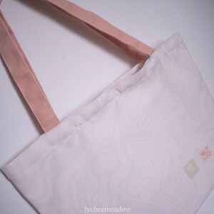 May include: A white tote bag with pink straps and a subtle marbled pattern. The bag has three small, light pink squares with a design in each.