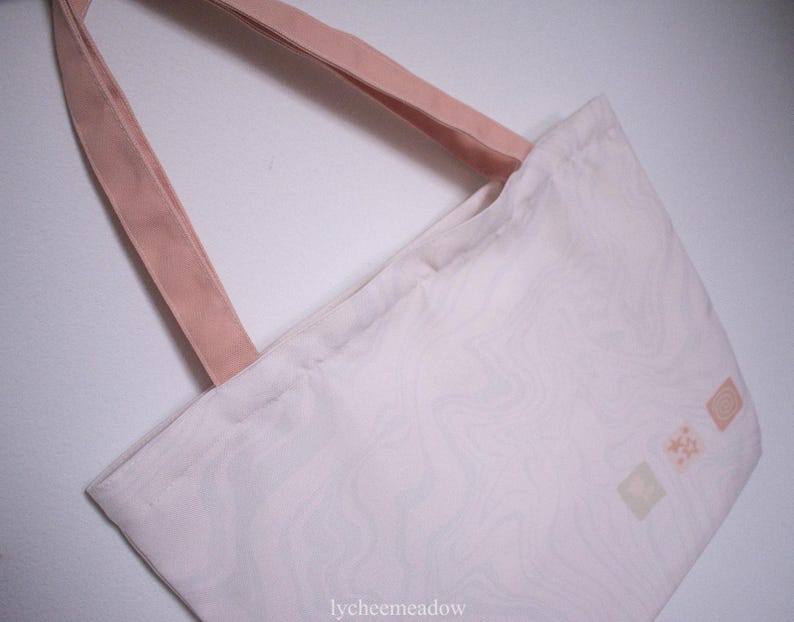 May include: A white tote bag with pink straps and a subtle marbled pattern. The bag has three small, light pink squares with a design in each.