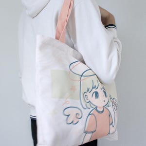 May include: A white tote bag with a pink strap and a cartoon illustration of a girl with angel wings and a halo. The girl is wearing a pink shirt and has blonde hair. The text "lycheemeadow" is printed on the bottom of the bag.