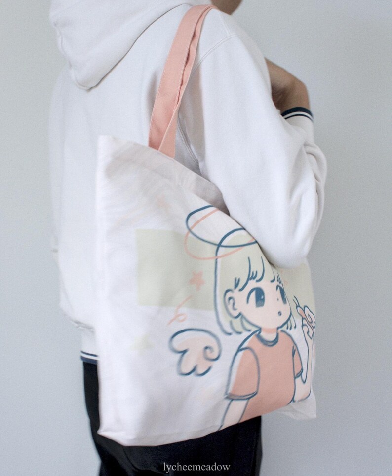 May include: A white tote bag with a pink strap and a cartoon illustration of a girl with angel wings and a halo. The girl is wearing a pink shirt and has blonde hair. The text "lycheemeadow" is printed on the bottom of the bag.