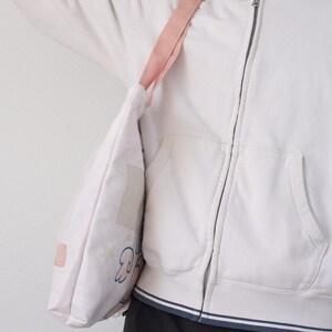 May include: A white tote bag with a pink strap and a cartoon design. The bag is being held by a person wearing a white hoodie.