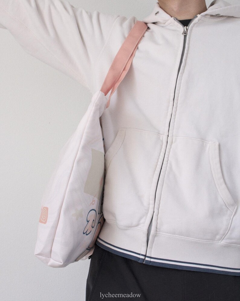 May include: A white tote bag with a pink strap and a cartoon design. The bag is being held by a person wearing a white hoodie.
