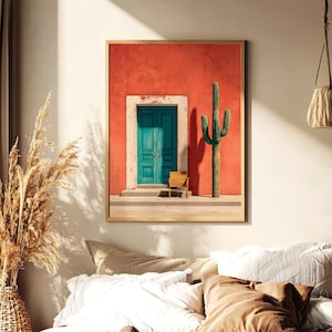 May include: A framed print featuring a vibrant scene: a teal door set in a red wall, with a cactus and small chair. The artwork has a warm, inviting aesthetic, with a touch of Southwestern charm. The print is encased in a simple, light-colored frame.