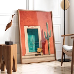 May include: A framed print featuring a vibrant red wall with a teal door and a cactus. A small wooden chair sits in front of the door. The print is leaning against a white wall. A wooden sculpture and a side table are on the left.