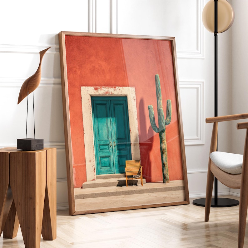 May include: A framed print featuring a vibrant red wall with a teal door and a cactus. A small wooden chair sits in front of the door. The print is leaning against a white wall. A wooden sculpture and a side table are on the left.