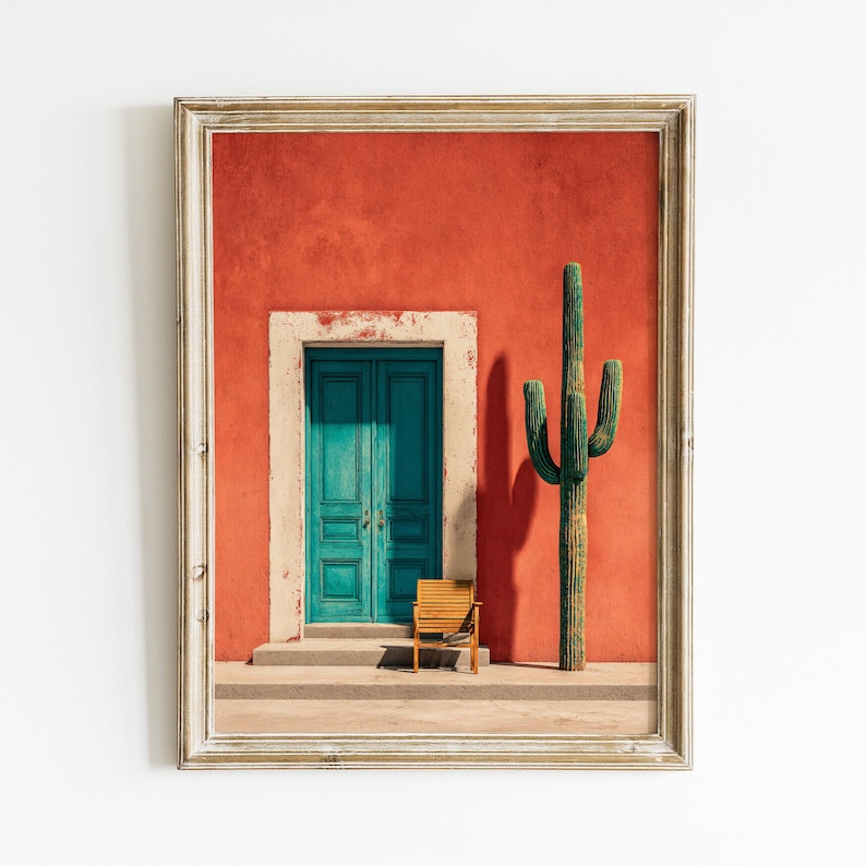 May include: Framed print featuring a teal door set in a red wall, with a tall green cactus and a small wooden chair. The image has a warm, desert-like aesthetic, with a neutral-toned frame.