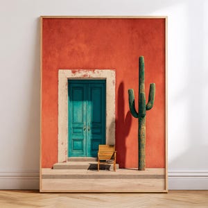 May include: A framed print featuring a vibrant scene with a teal door set in a red stucco wall. A tall green cactus stands beside the door, with a small wooden chair in front. The artwork is framed in a light wood.
