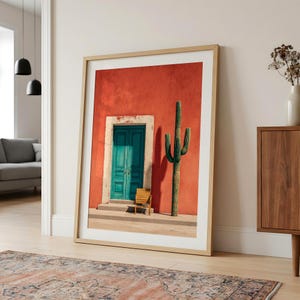 May include: Framed art print featuring a vibrant Southwestern scene. A teal door and a small wooden chair sit in front of a red wall, with a tall cactus to the right. The print is in a light wood frame.