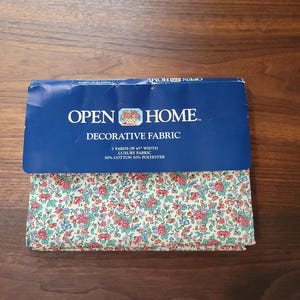 May include: A folded piece of decorative fabric with a floral pattern of small red, pink, and blue flowers on a cream background. The blue packaging label reads OPEN HOME DECORATIVE FABRIC, 3 YARDS OF 45 INCH WIDTH, 50 PERCENT COTTON 50 PERCENT POLYESTER.