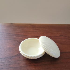 May include: A small, off-white ceramic trinket dish with a matching lid. The dish is round with a beaded edge, and the lid has a shell-like design. The dish and lid are resting on a dark wood surface.