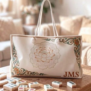 May include: A cream-colored canvas tote bag featuring a central lotus flower line drawing, ornate floral corner accents in muted green and orange, and the letters JMS printed in the bottom right corner, accompanied by scattered mahjong tiles.