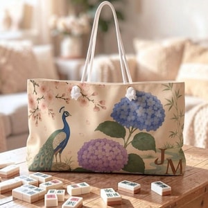 May include: A beige tote bag with white rope handles featuring a printed design of a peacock, pink cherry blossoms, and blue and purple hydrangea flowers. The bag includes the monogram letters JM in the bottom right corner of the exterior.