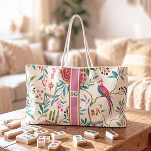 May include: A white tote bag featuring a colourful botanical print with pink birds, green leaves, and mahjong tiles. A vertical pink stripe runs down the centre with the initials SG printed in black. The bag has two white shoulder straps.