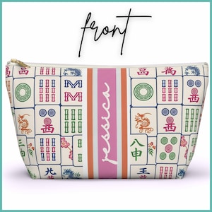 May include: A cream-coloured cosmetic bag with a gold zip, decorated with a pattern of colourful Mahjong tiles. A pink vertical stripe with the name "Jessica" in white script is centred on the bag.