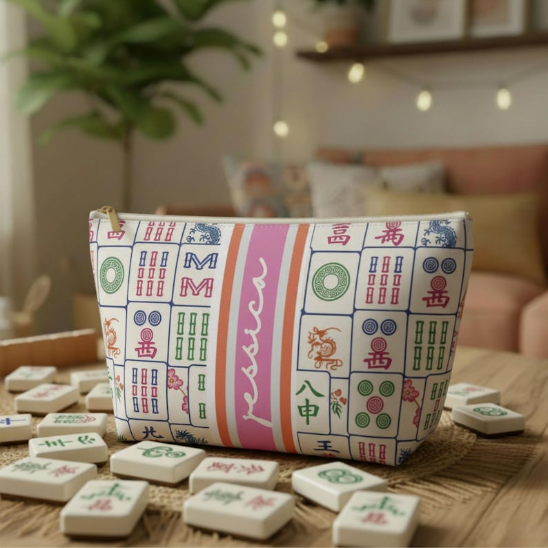 May include: A white cosmetic bag with a zip, decorated with Mahjong tiles in green, blue, and red. A pink stripe with the name "Jessica" is down the middle. Mahjong tiles are scattered on the table in front.