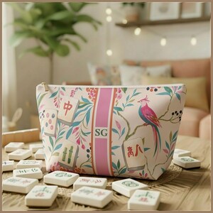 May include: A cream-colored zippered pouch featuring a botanical pattern of leaves, flowers, and a pink bird. The design includes scattered mahjong tiles and a central vertical pink stripe with the letters SG printed in a dark serif font.