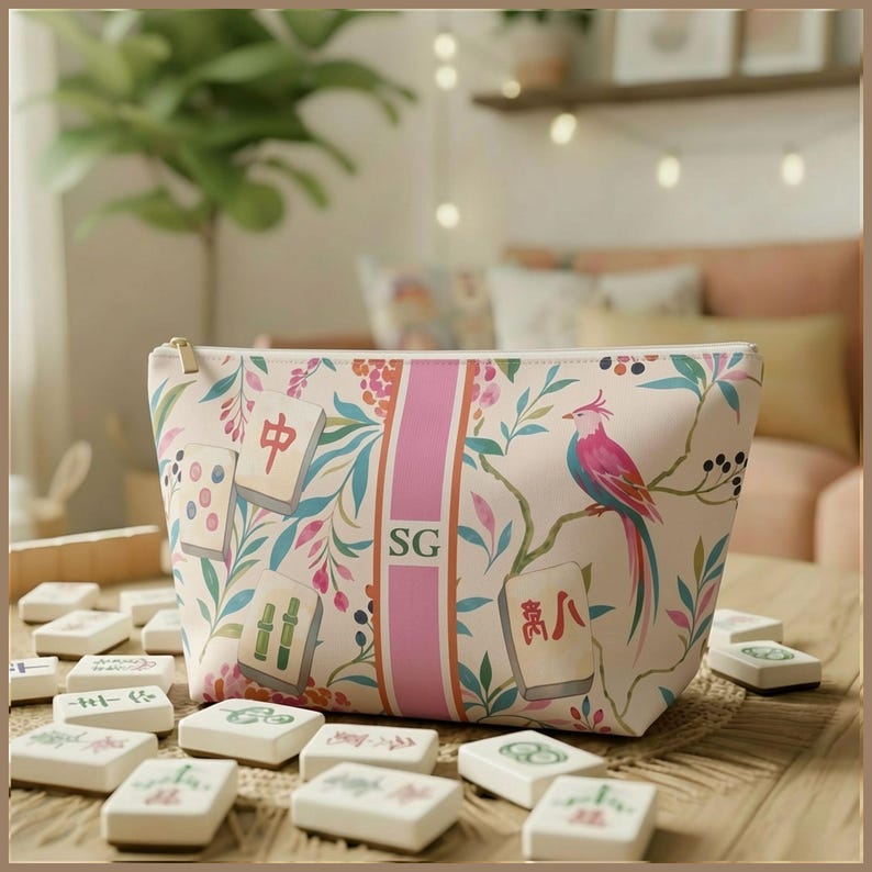 May include: A cream-colored zippered pouch featuring a botanical pattern of leaves, flowers, and a pink bird. The design includes scattered mahjong tiles and a central vertical pink stripe with the letters SG printed in a dark serif font.