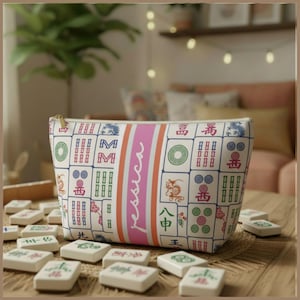 May include: A white zippered pouch featuring a colorful mahjong tile pattern and a central vertical pink stripe with the name Jessica printed in white script. The pouch is surrounded by scattered white mahjong tiles on a wooden surface.